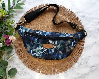 May include: A blue and white floral fanny pack with a black adjustable strap and gold hardware. The fanny pack is on a brown woven mat.