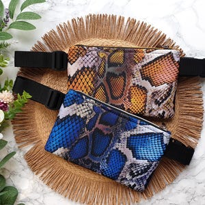 Op de afbeelding: Twee fanny packs met slangenprint en verstelbare zwarte banden. De bovenste tas is oranje en bruin, de onderste tas is blauw en wit.