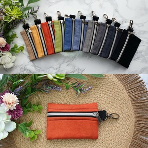 Könnte beinhalten: Eine Sammlung kleiner Reißverschlusstaschen in verschiedenen Farben, darunter Beige, Orange, Grün, Blau, Grau und Schwarz. Jede Tasche hat einen silbernen Reißverschluss und einen kleinen Metallverschluss. Eine orangefarbene Tasche wird separat gezeigt. Die Taschen werden auf einer Marmoroberfläche und einer gewebten Matte präsentiert.