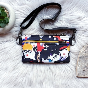 May include: A black crossbody bag with a colorful print of women's faces. The bag has two zippered compartments and a black adjustable strap.