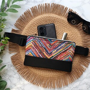 May include: A colorful fanny pack with a black strap, a black phone, and a silver tube are inside the pack. The pack is on a woven straw mat.