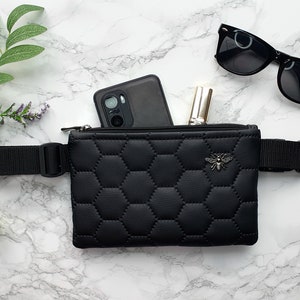 Può includere: Una marsupio nera trapuntata con una chiusura a zip e una tracolla nera regolabile. Il marsupio ha un design a forma di ape sul davanti ed è mostrato con uno smartphone e un rossetto all'interno.