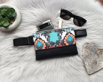 May include: A black fanny pack with a white, orange, and blue patterned front. The fanny pack has a black strap and is lying on a white fluffy surface. A cell phone, sunglasses, and a lipstick are inside the fanny pack.