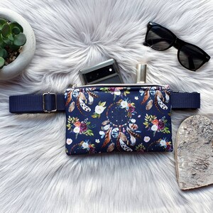 Könnte beinhalten: Eine marineblaue Bauchtasche mit einem Blumen- und Federmuster. Die Tasche hat einen Reißverschluss und einen verstellbaren Gurt. Die Tasche liegt auf einer weißen, flauschigen Oberfläche mit einem Telefon, einer Sonnenbrille und einem Holzhertz.