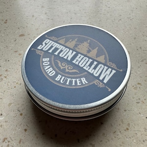 May include: A silver tin with a blue label that reads "Sutton Hollow Board Butter". The label features a stylized design of trees and a decorative border.