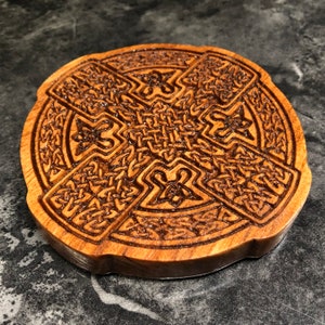 May include: A round wooden coaster with a Celtic knot design carved into the surface. The coaster is made of dark wood and has a natural finish.