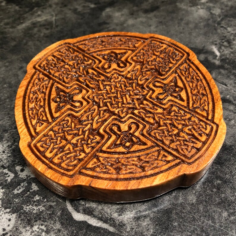 May include: A round wooden coaster with a Celtic knot design carved into the surface. The coaster is made of dark wood and has a natural finish.