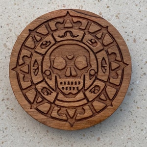 May include: A wooden coaster with a carved skull and crossbones design. The skull is surrounded by a circle of intricate carvings.