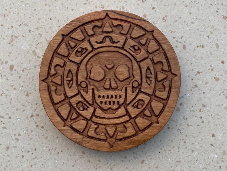 May include: A wooden coaster with a carved skull and crossbones design. The skull is surrounded by a circle of intricate carvings.