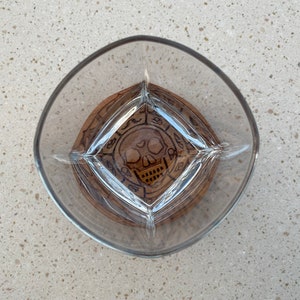 May include: A clear glass with a wooden coaster featuring a skull and crossbones design. The coaster has a dark brown wood finish and a circular shape.