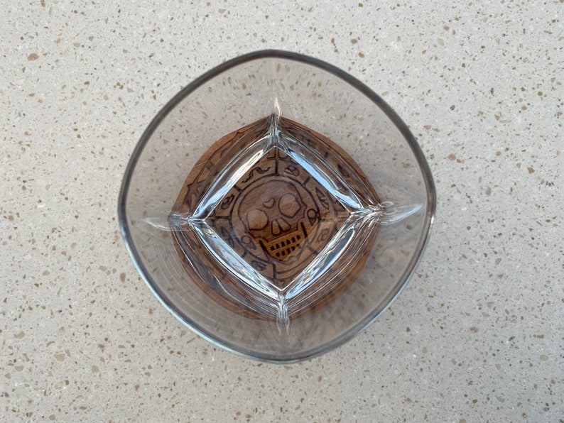 May include: A clear glass with a wooden coaster featuring a skull and crossbones design. The coaster has a dark brown wood finish and a circular shape.