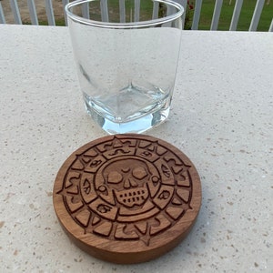 May include: A wooden coaster with a carved skull and compass design. The coaster is brown and has a circular shape.