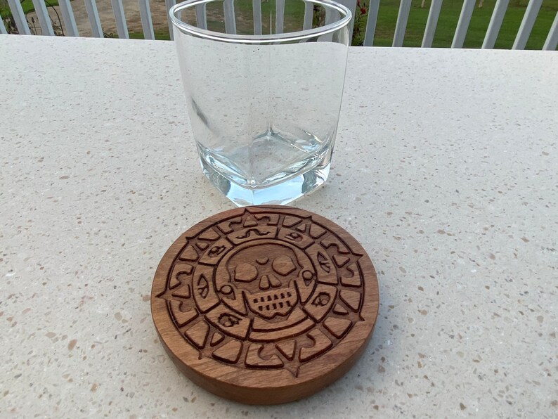 May include: A wooden coaster with a carved skull and compass design. The coaster is brown and has a circular shape.