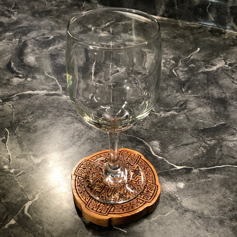 May include: A clear glass wine glass sits on a wooden coaster with a Celtic knot design. The coaster is brown and has a detailed intricate pattern.