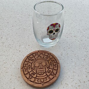 May include: A wooden coaster with a carved design of a skull and a circle of symbols. A clear glass with a sugar skull design is sitting on top of the coaster.