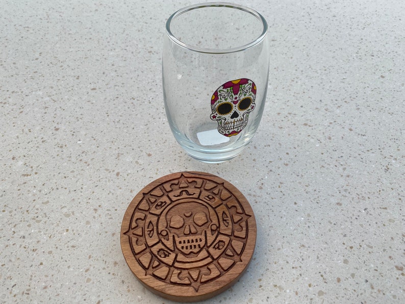 May include: A wooden coaster with a carved design of a skull and a circle of symbols. A clear glass with a sugar skull design is sitting on top of the coaster.