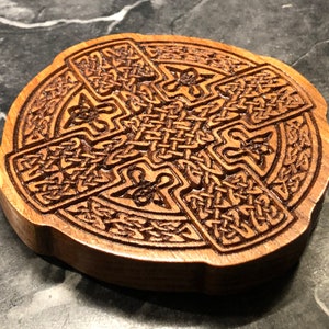May include: A wooden coaster with a Celtic knot design carved into the surface. The coaster is circular and has a brown wood finish.