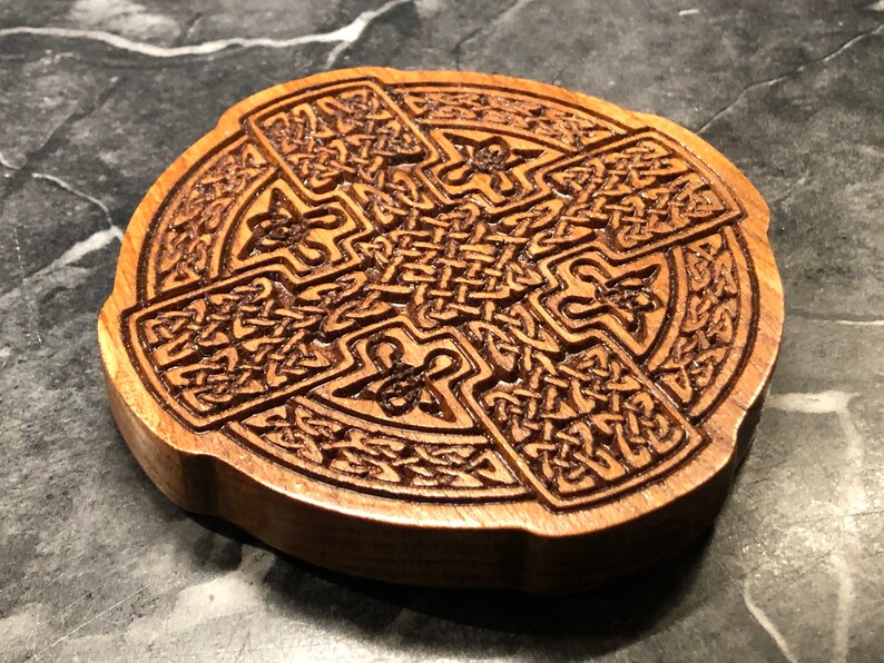 May include: A wooden coaster with a Celtic knot design carved into the surface. The coaster is circular and has a brown wood finish.