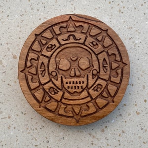 May include: A wooden coaster with a carved skull and crossbones design. The skull has a heart shape in the center of its forehead.