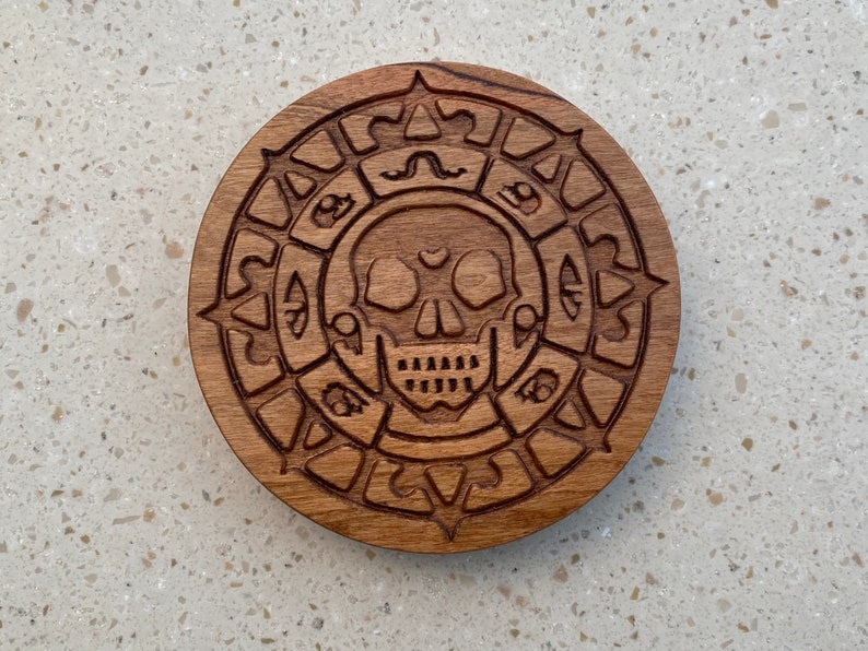 May include: A wooden coaster with a carved skull and crossbones design. The skull has a heart shape in the center of its forehead.