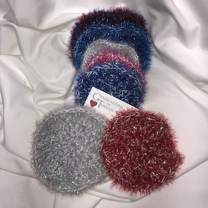 May include: A set of six colorful crocheted dishcloths with a sparkly finish. The dishcloths are in shades of red, blue, pink, and gray. The dishcloths are stacked on top of each other and are labeled "Generations of Thread".