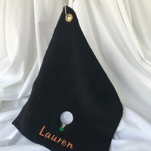 May include: Black golf towel with a white golf ball and green grass embroidered on it. The towel has a gold metal ring and the name "Lauren" is embroidered in orange thread.