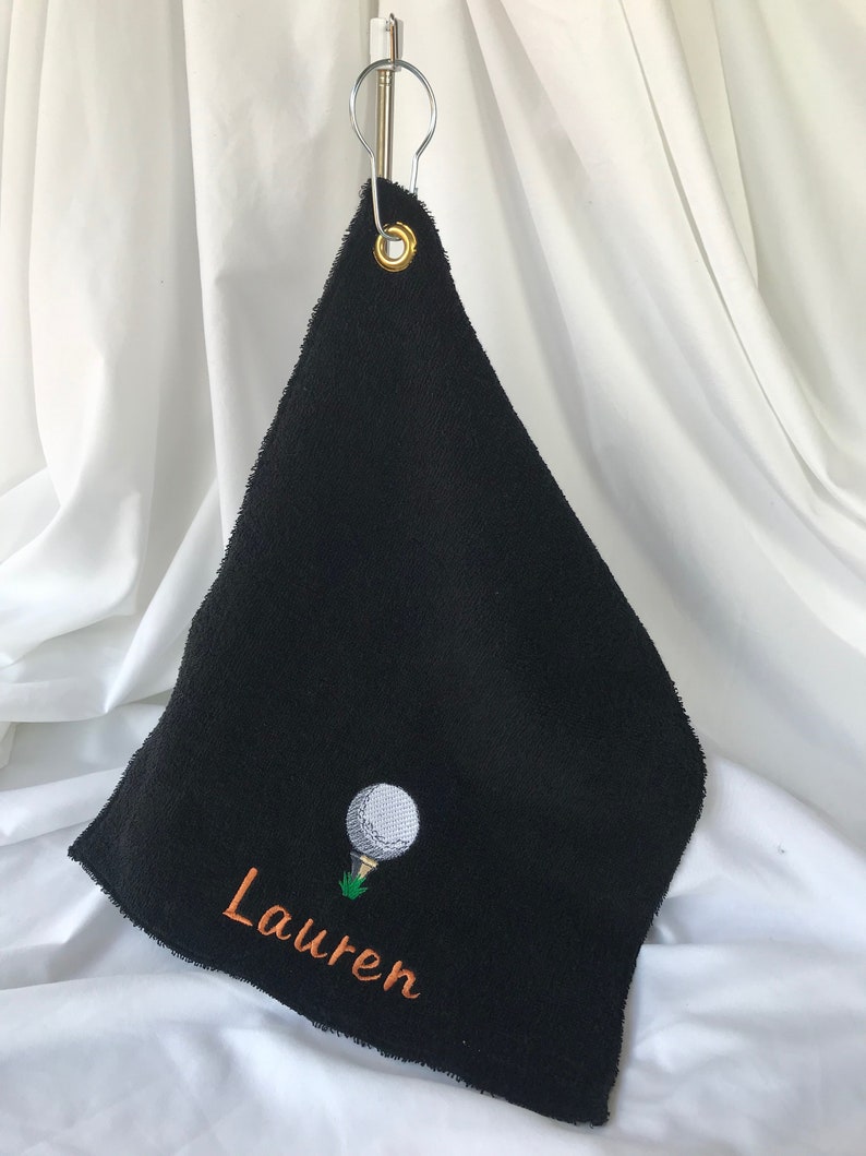 May include: Black golf towel with a white golf ball and green grass embroidered on it. The towel has a gold metal ring and the name "Lauren" is embroidered in orange thread.