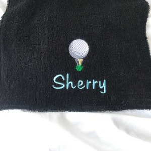 May include: Black golf towel with a white golf ball and green grass embroidered on it. The name "Sherry" is embroidered in blue below the golf ball.