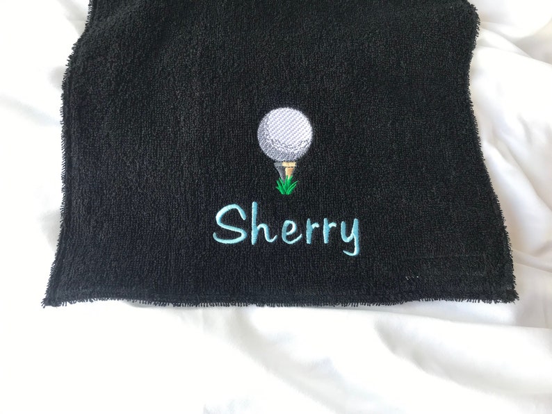 May include: Black golf towel with a white golf ball and green grass embroidered on it. The name "Sherry" is embroidered in blue below the golf ball.