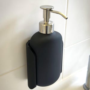 Pode incluir: Um dispensador de sabonete preto fosco com bomba e bico prateados. O dispensador é montado em uma parede de azulejos brancos. O dispensador é mantido no lugar por um suporte preto.