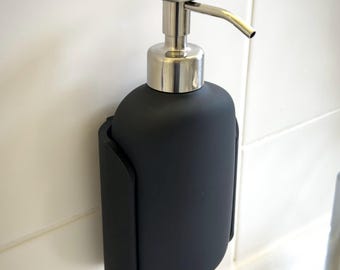Könnte beinhalten: Ein mattschwarzer Seifenspender mit silberner Pumpe und Ausguss. Der Spender ist an einer weiß gefliesten Wand montiert. Der Spender wird von einem schwarzen Halter gehalten.