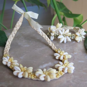 May include: A natural-toned necklace and bracelet set. The necklace and bracelet are adorned with white seashells and yellow accents, and the necklace has a braided band with a cream-colored ribbon tie.