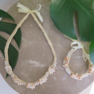 May include: A woven necklace and matching bracelet, both adorned with seashells. The necklace and bracelet are made of a light beige woven material and are decorated with small, light-colored seashells. A light yellow ribbon is tied at the top of each piece.