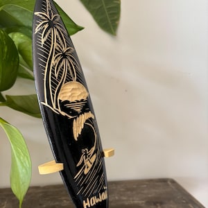 May include: A black wooden surfboard with a gold carving of palm trees, a surfer, and a wave. The surfboard is on a wooden stand and says "Hawaii" at the bottom.