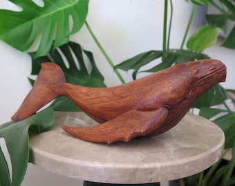 May include: A handcrafted wooden whale sculpture with a rich brown finish. The whale features detailed carving, including fins and a tail. The sculpture is displayed on a round, light-colored surface, with green foliage in the background.