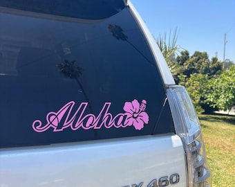 May include: A pink vinyl decal on the rear window of a white vehicle. The decal displays the word "Aloha" in a cursive font, alongside a pink hibiscus flower. The vehicle model is a GX 460.