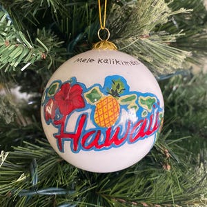 May include: White Christmas ornament with a painted image of the Hawaiian islands, a pineapple, a hibiscus flower, and the text "Mele Kalikimaka Hawaii".