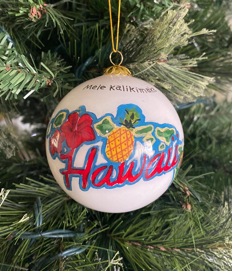May include: White Christmas ornament with a painted image of the Hawaiian islands, a pineapple, a hibiscus flower, and the text "Mele Kalikimaka Hawaii".