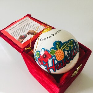 May include: White ceramic Christmas ornament with a hand-painted design of a pineapple, hibiscus flower, and the word "Hawaii" in blue and red lettering. The ornament is in a red velvet box with a certificate of authenticity that reads "Hand painted from the inside" and "Mele Kalikimaka".