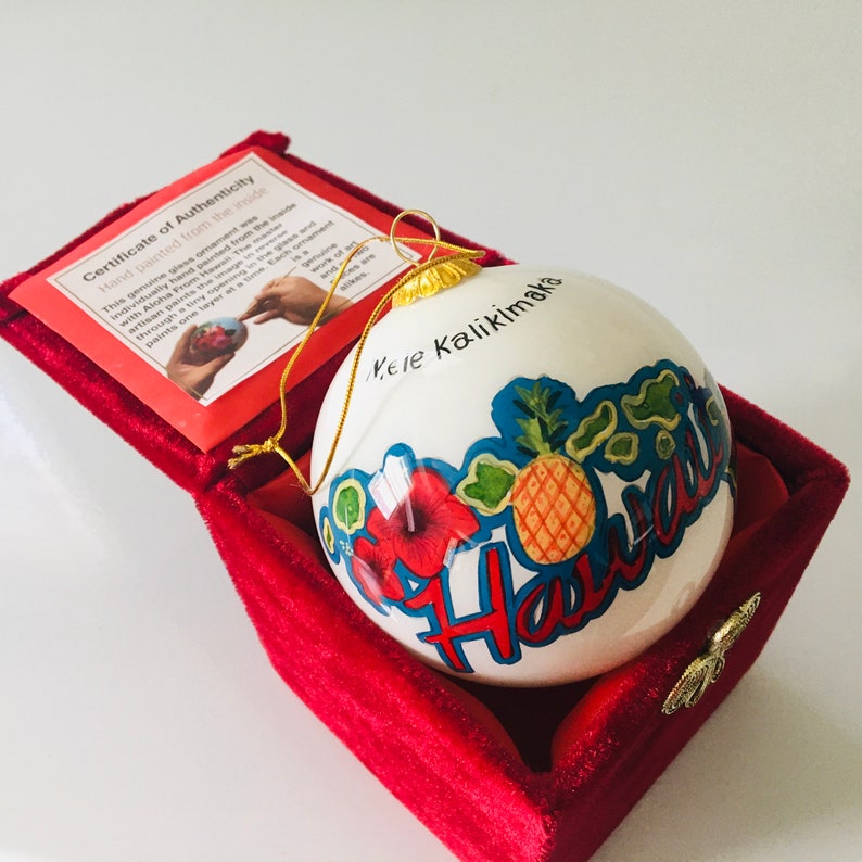 May include: White ceramic Christmas ornament with a hand-painted design of a pineapple, hibiscus flower, and the word "Hawaii" in blue and red lettering. The ornament is in a red velvet box with a certificate of authenticity that reads "Hand painted from the inside" and "Mele Kalikimaka".