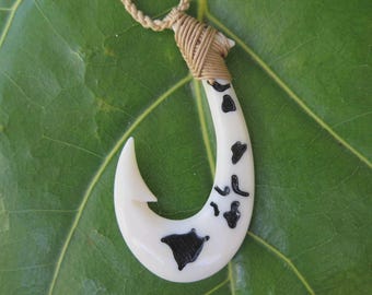 May include: Cream-colored, carved bone pendant in the shape of a fish hook, with black markings. The pendant is suspended from a tan cord necklace. The background is a large green leaf.