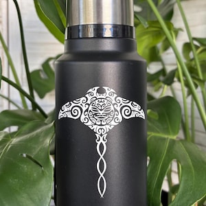 May include: Black insulated water bottle with a silver lid. The bottle features a white tribal design of a manta ray. The bottle is surrounded by green leafy plants.