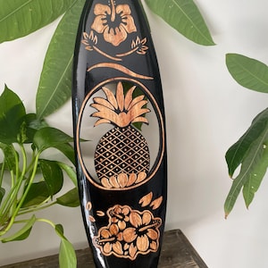 May include: Black wooden surfboard-shaped wall decoration with carved hibiscus flowers, a pineapple, and the word "HAWAII". The pineapple is in an oval cutout. The surfboard is surrounded by green leaves.
