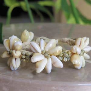 May include: A handcrafted bracelet featuring three flower-like clusters made of white and cream seashells. The clusters are connected by a woven, beige cord and accented with small, green beads. The bracelet rests on a light-colored surface.