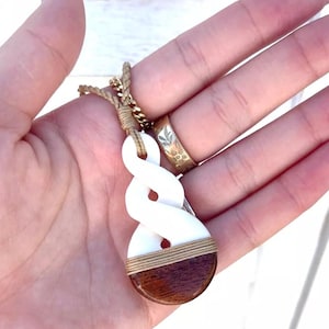 May include: A white bone pendant with a brown wood accent, carved in a twisted rope design. The pendant is attached to a brown cord necklace.