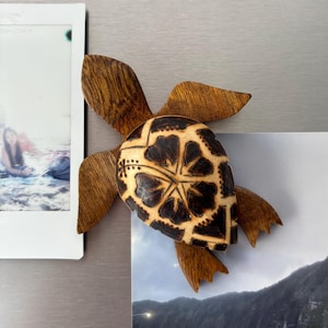 May include: Wooden turtle-shaped magnet with a detailed design. The turtle's shell features a light brown background with a dark brown floral pattern. The flippers and head are a darker brown. The magnet is attached to a refrigerator with a photo.