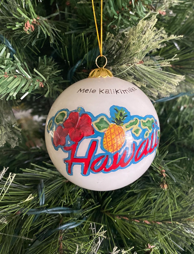May include: White Christmas ornament with a red hibiscus flower, a pineapple, and the words "Mele Kalikimaka Hawaii" in blue and red lettering.