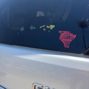 Puede incluir: En la ventana de un coche se exhiben pegatinas de vinilo coloridas de las islas hawaianas. Las pegatinas son de color verde, amarillo y rojo, cada una con intrincados diseños tribales. La carrocería plateada del coche y el emblema GX 460 son visibles.