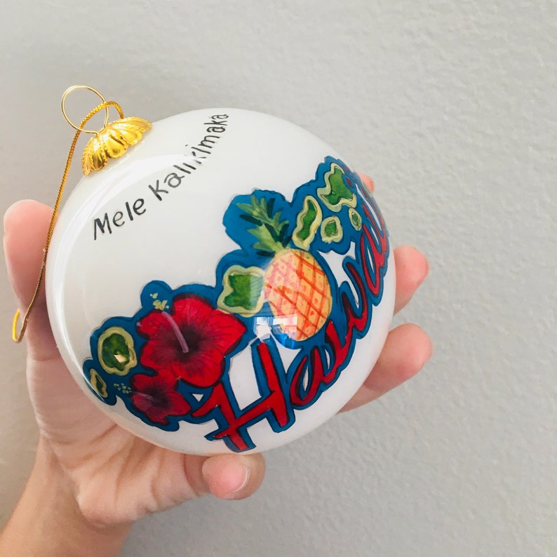 May include: White Christmas ornament with a pineapple, hibiscus flowers, and the text "Mele Kalikimaka Hawaii".