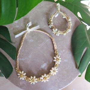 May include: A floral necklace and matching bracelet set. The necklace and bracelet are made of woven straw and adorned with white daisy-like flowers and small gold beads. The set is tied with a white ribbon.